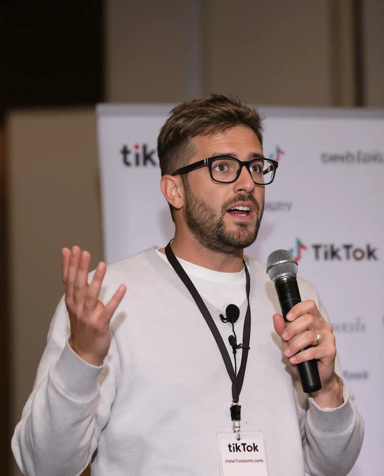 Flux.1 AI prompt A charismatic speaker is captured mid-speech. He has short, tousled brown hair that's slightly messy on top. He has a round circle face, clean shaven, adorned with rounded rectangular-framed glasses with dark rims, is animated as he gestures with his left hand. He is holding a black microphone in his right hand, speaking passionately.The man is wearing a light grey sweater over a white t-shirt. He's also wearing a simple black lanyard hanging around his neck. The lanyard badge has the text TikTok .Behind him, there is a blurred background with a white banner containing logos and text (including tiktok), a professional conference setting.